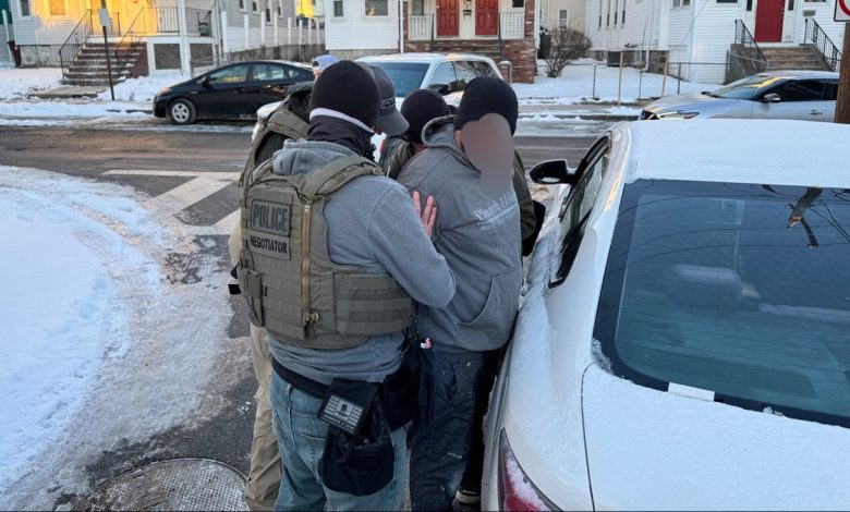 Three ICE officers stand around a man with a blurred out face as they put him in handcuffs.