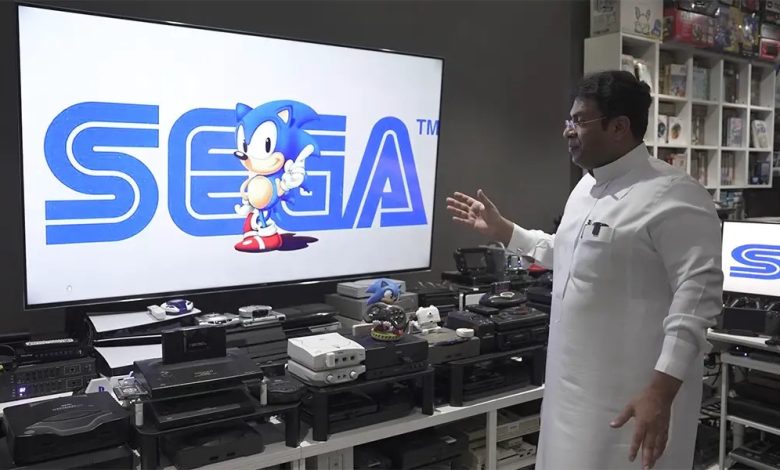 Ibrahim Al-Nasser standing in front of his console setup. There are a ton of consoles lined up in front of a TV. Sonic and the Sega logo are on the TV