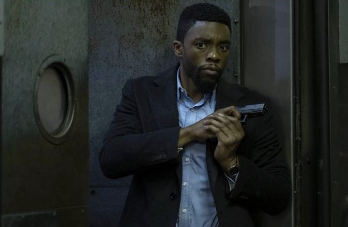 Chadwick Boseman holds a gun and stands up against a wall.