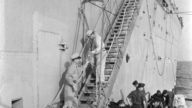 A sailor walking down staircase on side of ship to hand a jar of fluid to a sailor at the bottom, surrounded by other sailors