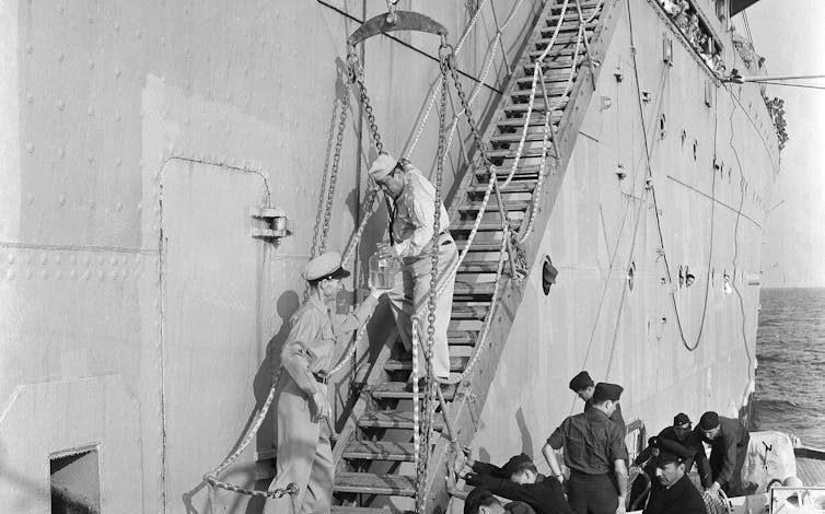 A sailor walking down staircase on side of ship to hand a jar of fluid to a sailor at the bottom, surrounded by other sailors