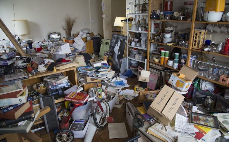 A room full of piles of papers, stacked shelves and a tricycle.