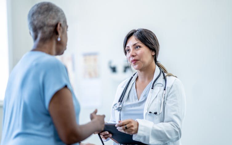 A gray-haired Black woman speaks with a doctor in scrubs and a stethoscope, who is seated in front of her.