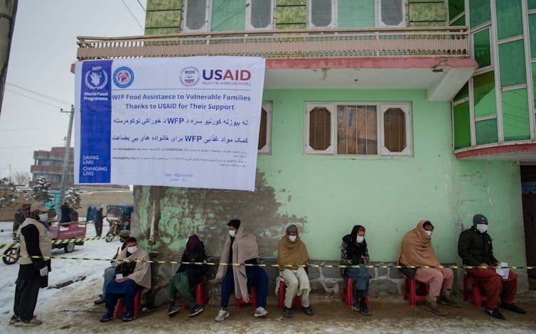 With a USAID banner hanging on a battered green building, a group of men sit outdoors, just below the banner.