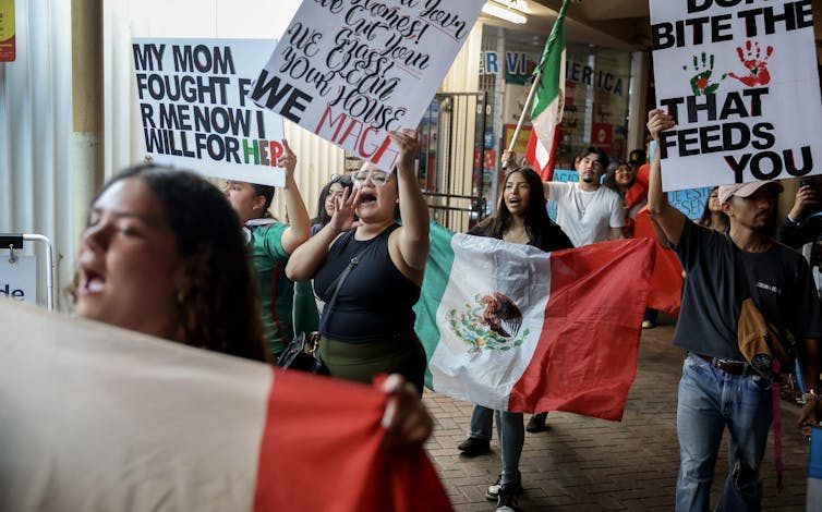 A group of protestors holding signs and yelling walk down the street. Some are holding Mexican flags.