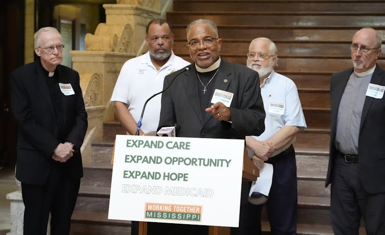 Religious men, some in collars, stand together around a sign calling for Medicaid expansion in Mississippi.