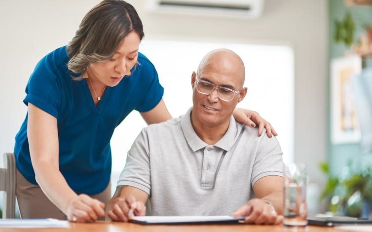 A woman in a blue shirt stands over a man who is sitting at a table filling out a document.