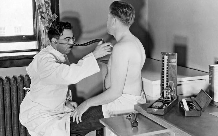 white-coated doctor uses stethoscope to listen to shirtless man's chest in exam room