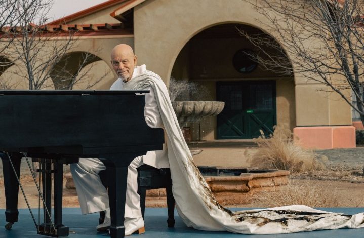 John Malkovich sits at a piano and smirks.