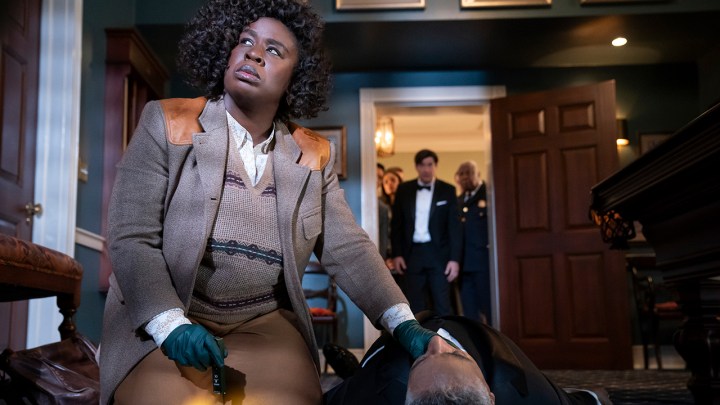 A woman with her hand on a man's neck who is lying dead on the ground as she looks up and others look on in the doorway behind her in The Residence.