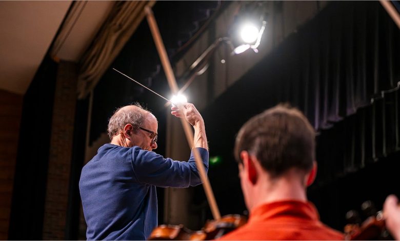 Rand Laycock pictured from behind as he leads an orchestra, wand in hand. Another person is pictured in the foreground.