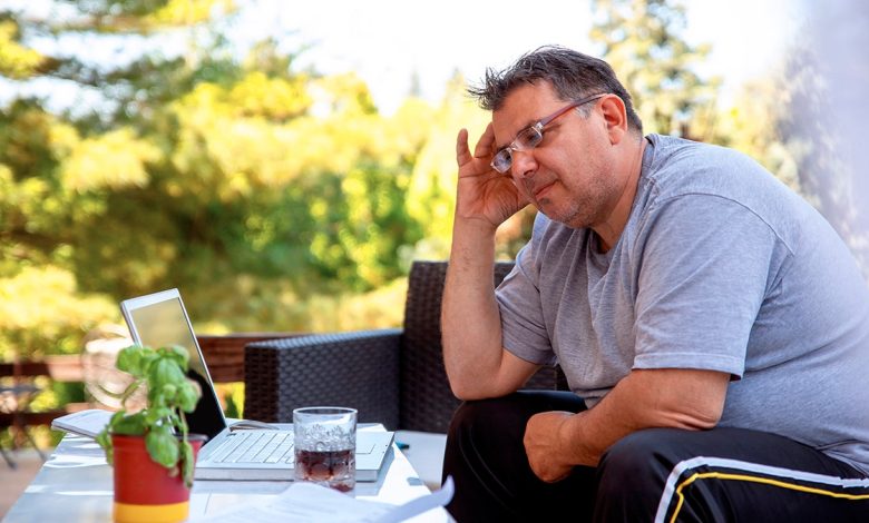 An overweight man sits outside on a chair, his hand pressed to his head. He looks disgruntled.