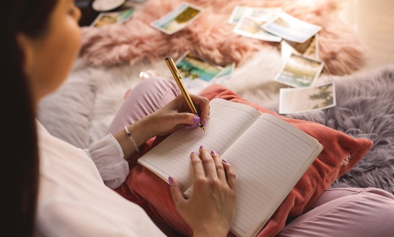 A young woman sits on her bed, writing in her journal with her left hand.