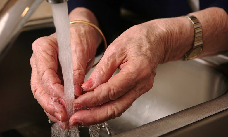 A pair of hands belonging to an older woman. The hands are being washed under a faucet in a sink.