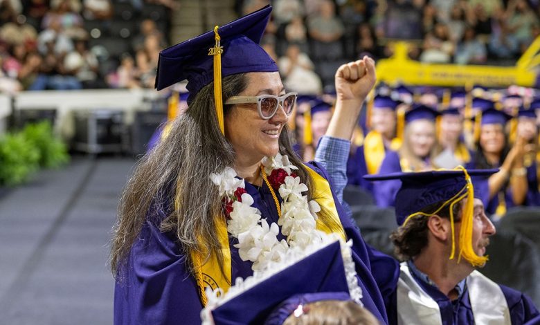 A blind graduate sits in her cap and gown at commencement with her guide dog resting at her feet.