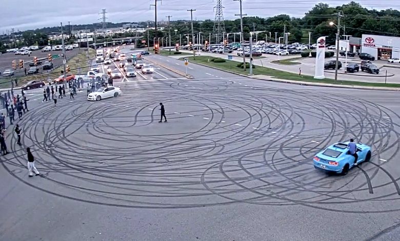 a large crowd surrounds intersection during street takover as cars make multiple donut marks on street