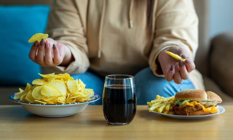 Woman sitting on couch with chips soda and burger in front of her.