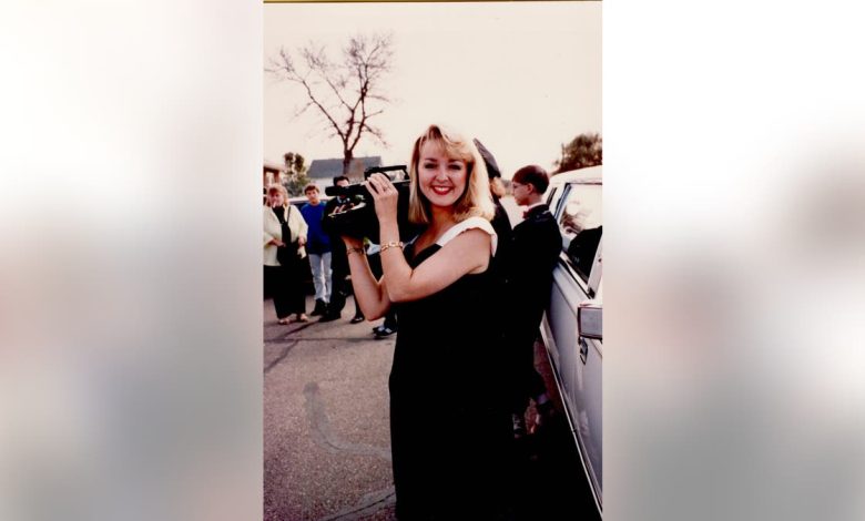 Jodi Huisentruit holding a camera wearing a black dress and smiling.